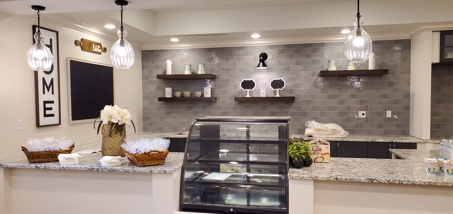 Interior view of a kitchen or serving area with granite countertops, two hanging pendant lights, a display case in the center, shelves with cups and pitchers on a gray tiled backsplash, and baskets with wrapped items on the counter. A decorative sign with the word 'HOME' is on the left wall.