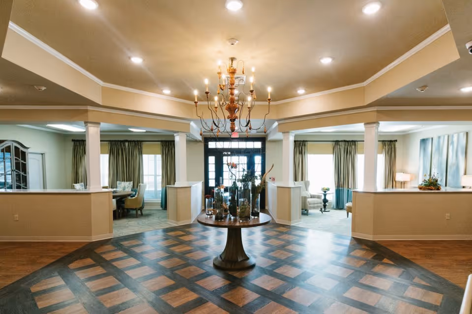 Spacious and well-lit interior common area with a central round table decorated with vases and plants, a chandelier hanging from the ceiling, and seating areas with chairs and tables visible through open partitions on either side.