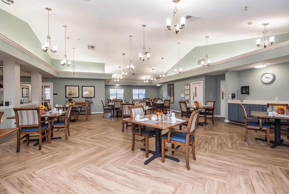 A spacious dining room in a senior living facility with multiple wooden tables and chairs arranged neatly. Each table is set with white cups, glasses, and small flower arrangements. The room has light green walls, large windows allowing natural light, and multiple ceiling light fixtures. There is a clock on the wall and a counter with a sink and paper towel dispenser.