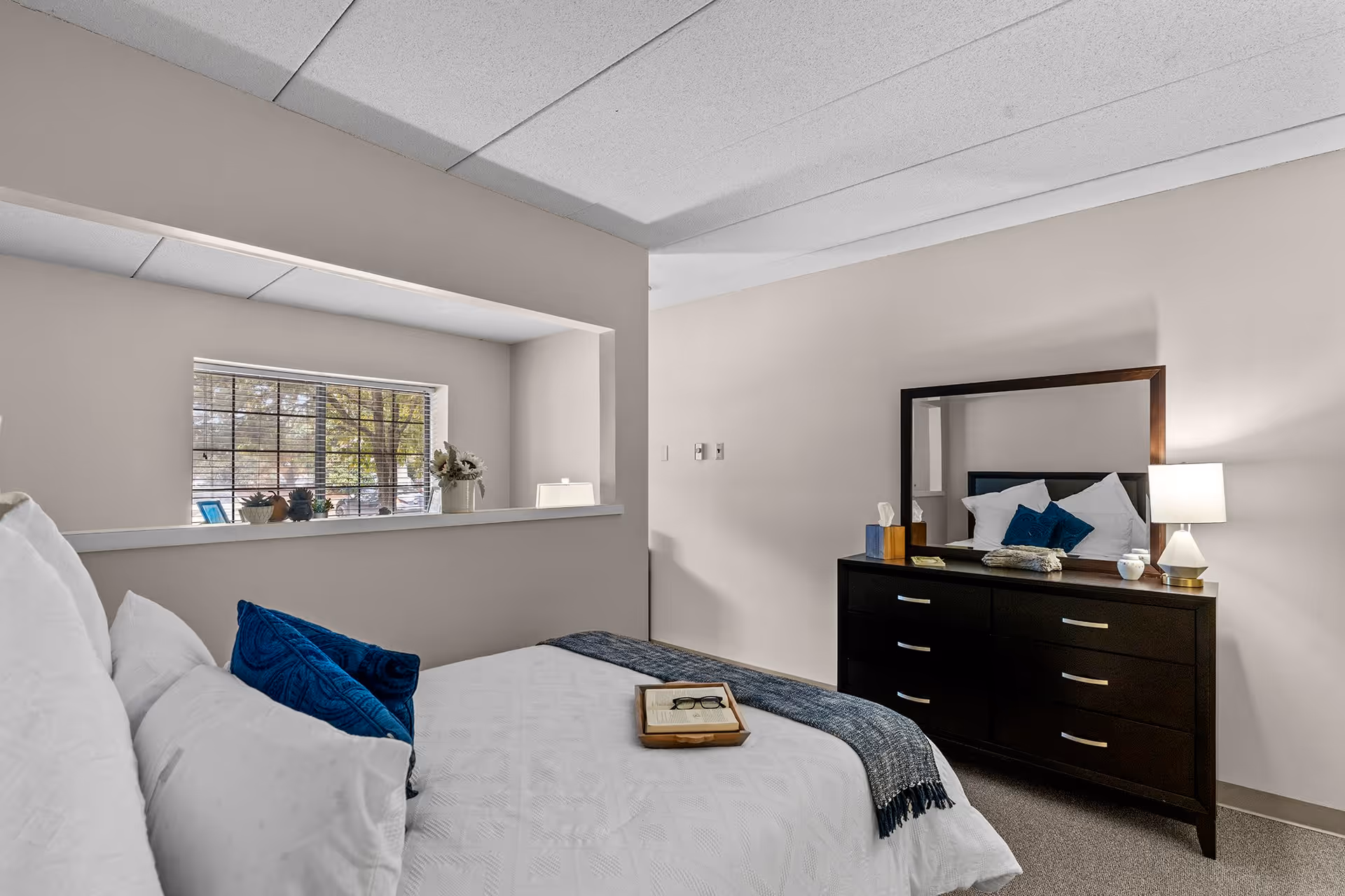 A bedroom with a neatly made bed featuring white bedding and blue accent pillows. A wooden dresser with a large mirror stands against the wall, topped with a lamp, tissue box, and decorative items. A window with blinds lets in natural light, and a small ledge below the window holds plants and a vase with flowers.
