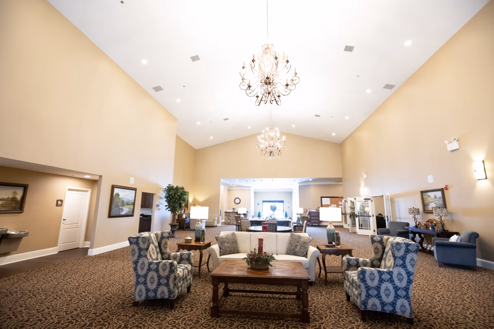 Spacious senior living common room with a vaulted ceiling and chandeliers, furnished with sofas, patterned armchairs, coffee tables, and lamps.
