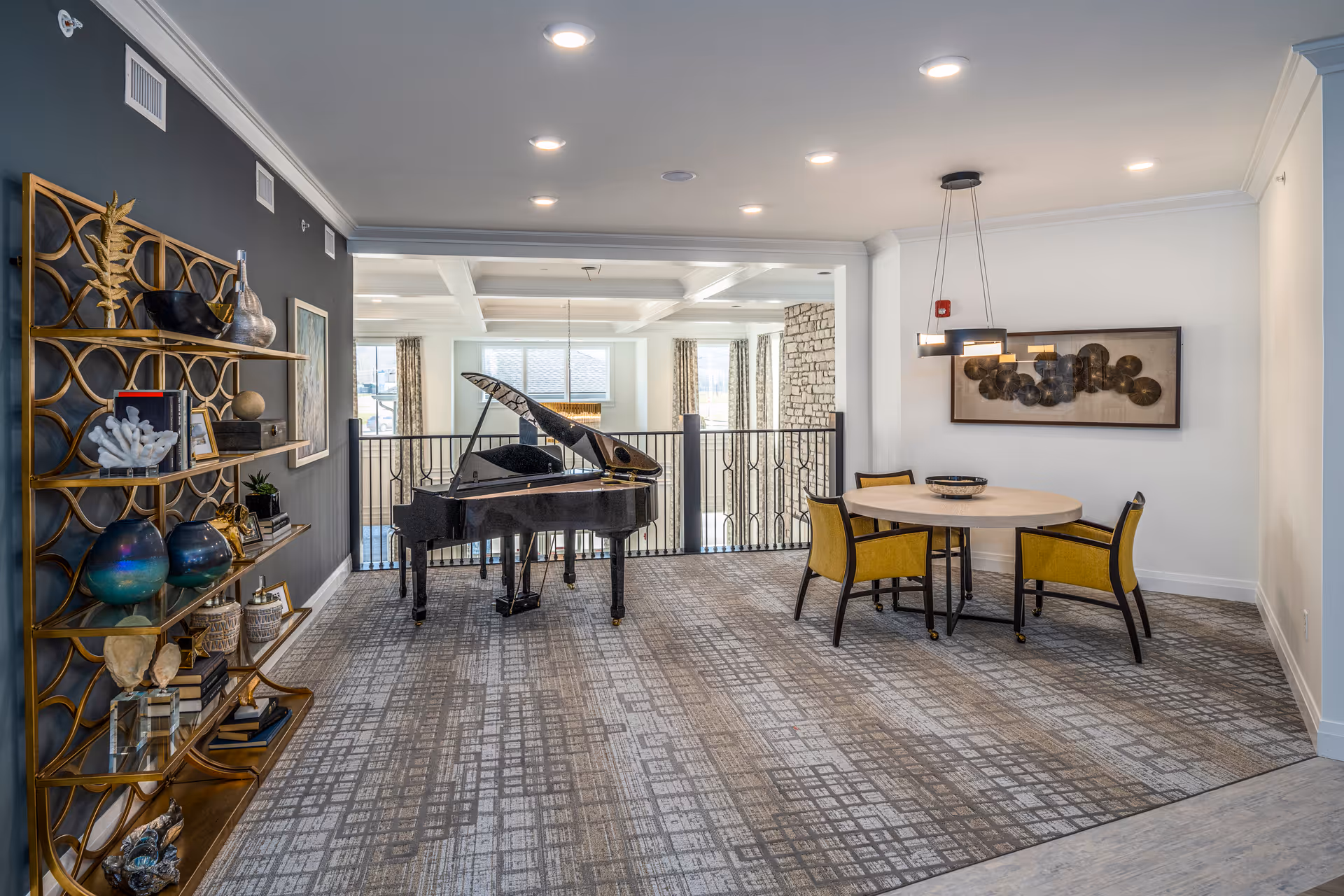 A spacious, well-lit interior room featuring a black grand piano near a railing overlooking a lower level. To the right, there is a round table with four yellow cushioned chairs and a modern hanging light fixture above. On the left side, a decorative gold shelving unit holds various art pieces and decor items. The room has a patterned carpet and white walls with some artwork.