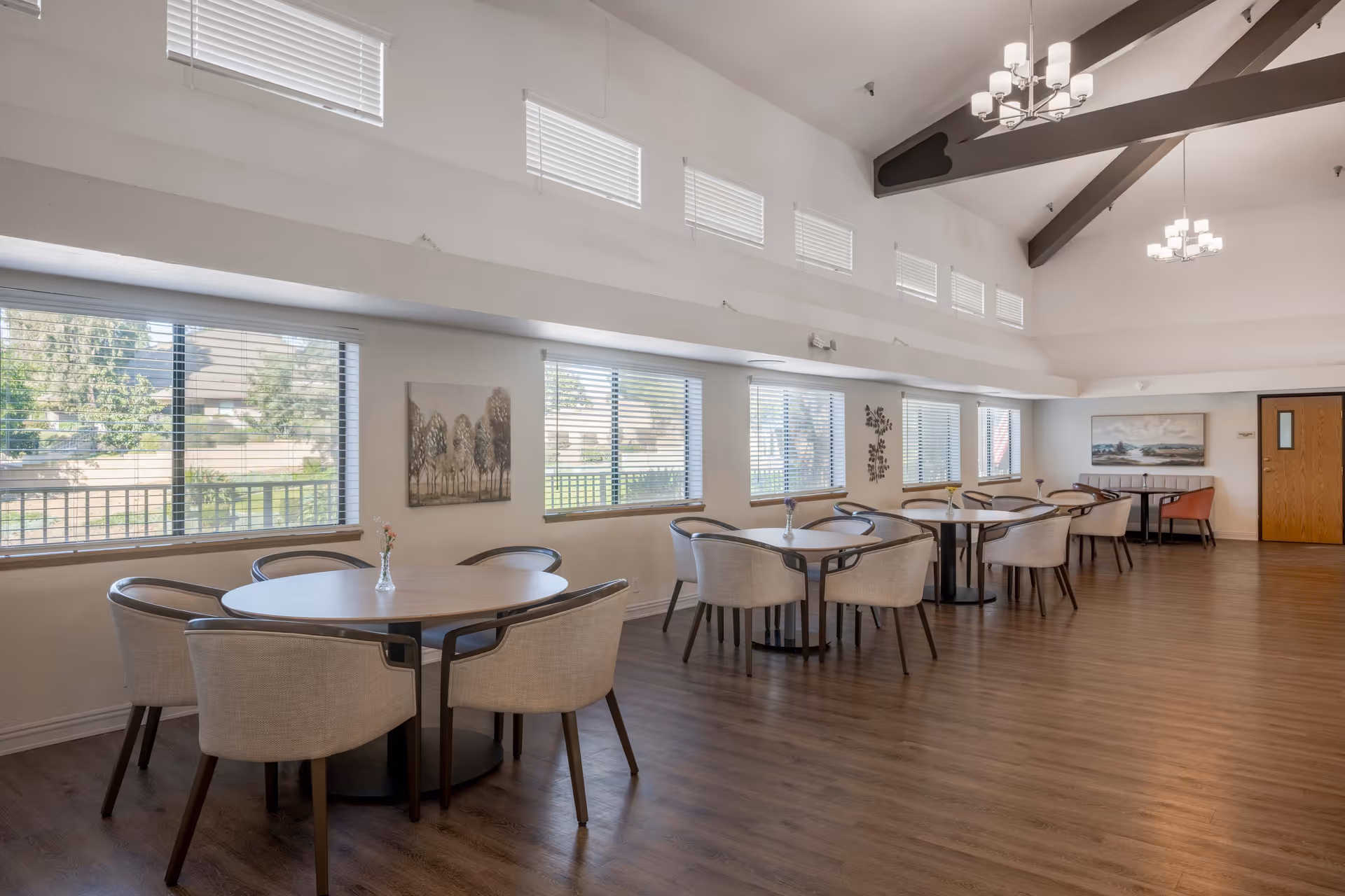 A spacious dining room with multiple round tables, each surrounded by four beige upholstered chairs. The room has large windows with blinds allowing natural light to enter, wooden flooring, and high ceilings with exposed beams and modern chandeliers. There are decorative wall art pieces and a door at the far end of the room.