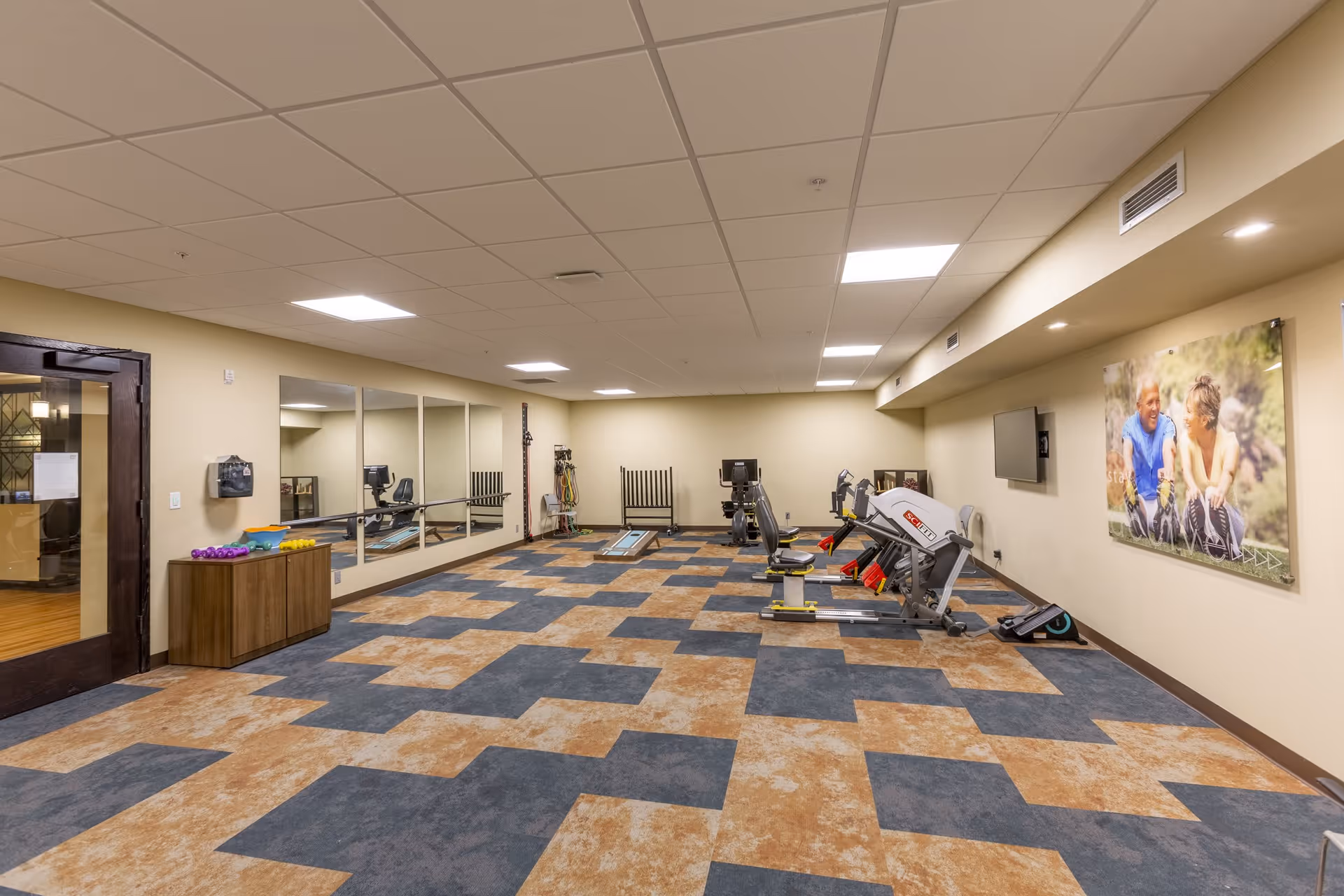 A spacious exercise room with patterned carpet flooring, several pieces of fitness equipment including a rowing machine and a leg press, a wall-mounted TV, large mirrors on one wall, and a colorful photograph of two people stretching on another wall. There is a wooden cabinet with small dumbbells and exercise bands near the entrance door.