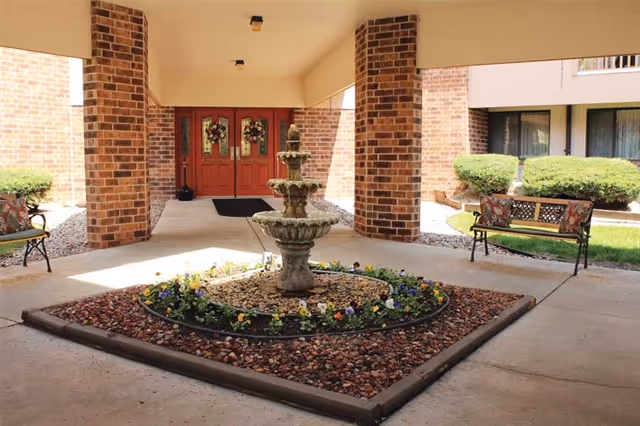 Covered outdoor entrance area with a three-tiered stone fountain surrounded by flowers and rocks in the center. There are brick pillars supporting the roof, two benches with floral cushions on either side, and double red doors decorated with wreaths in the background.
