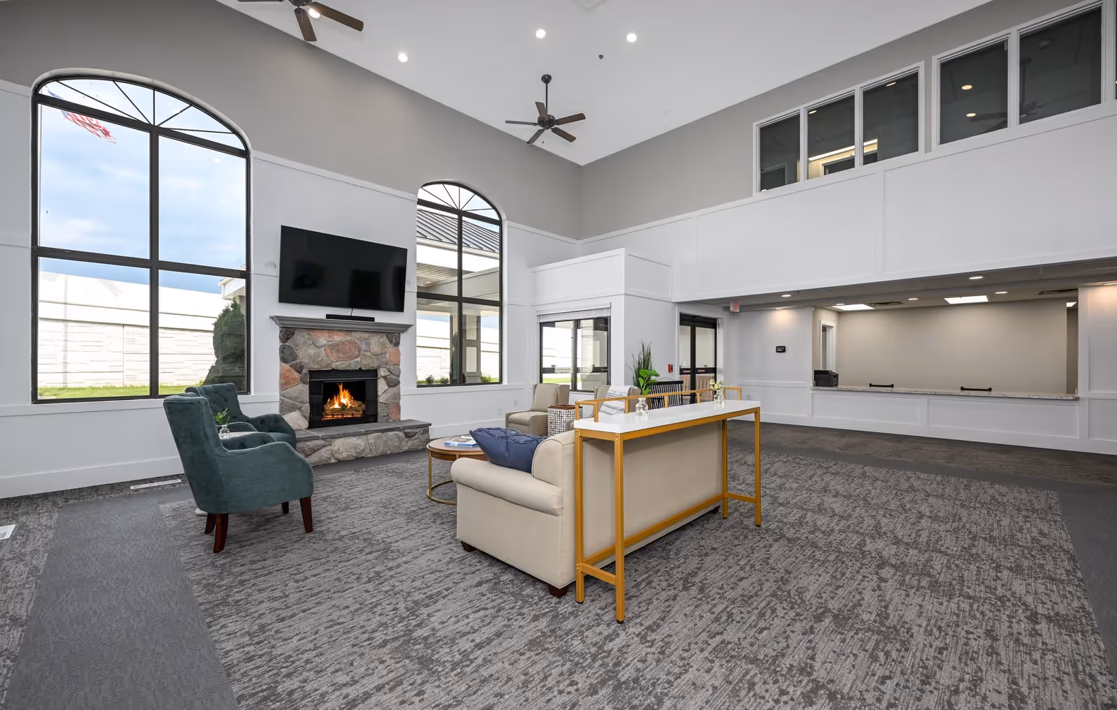 A spacious senior living facility common area with high ceilings, large arched windows, a stone fireplace with a fire burning, a wall-mounted flat-screen TV, and comfortable seating including armchairs and sofas arranged around a coffee table. The room has ceiling fans and a reception or service counter area in the background.
