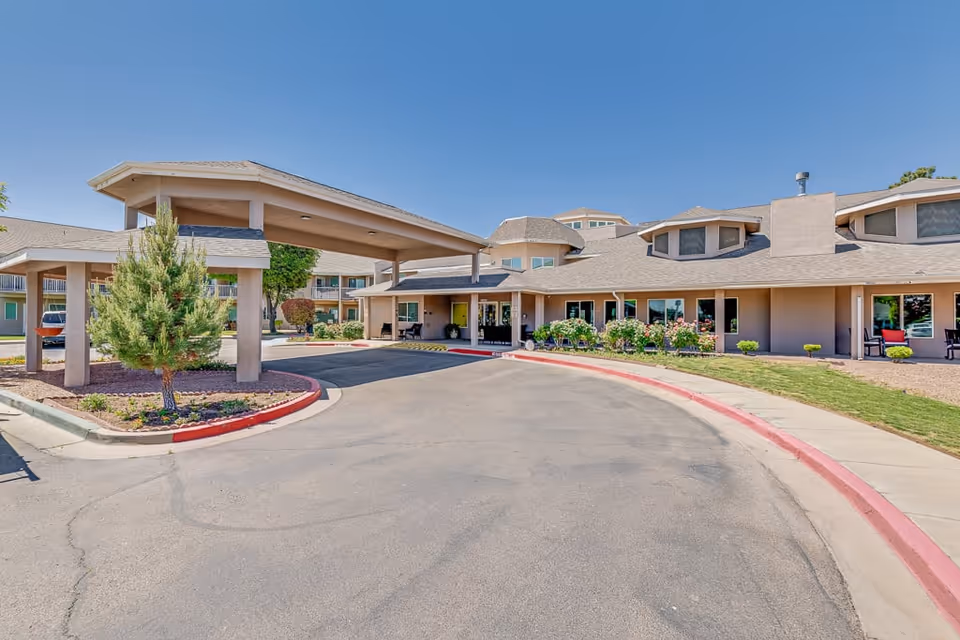 Exterior view of Solstice Senior Living at Rio Norte showing the main entrance with a covered drop-off area, surrounded by landscaping including small trees and bushes, under a clear blue sky.