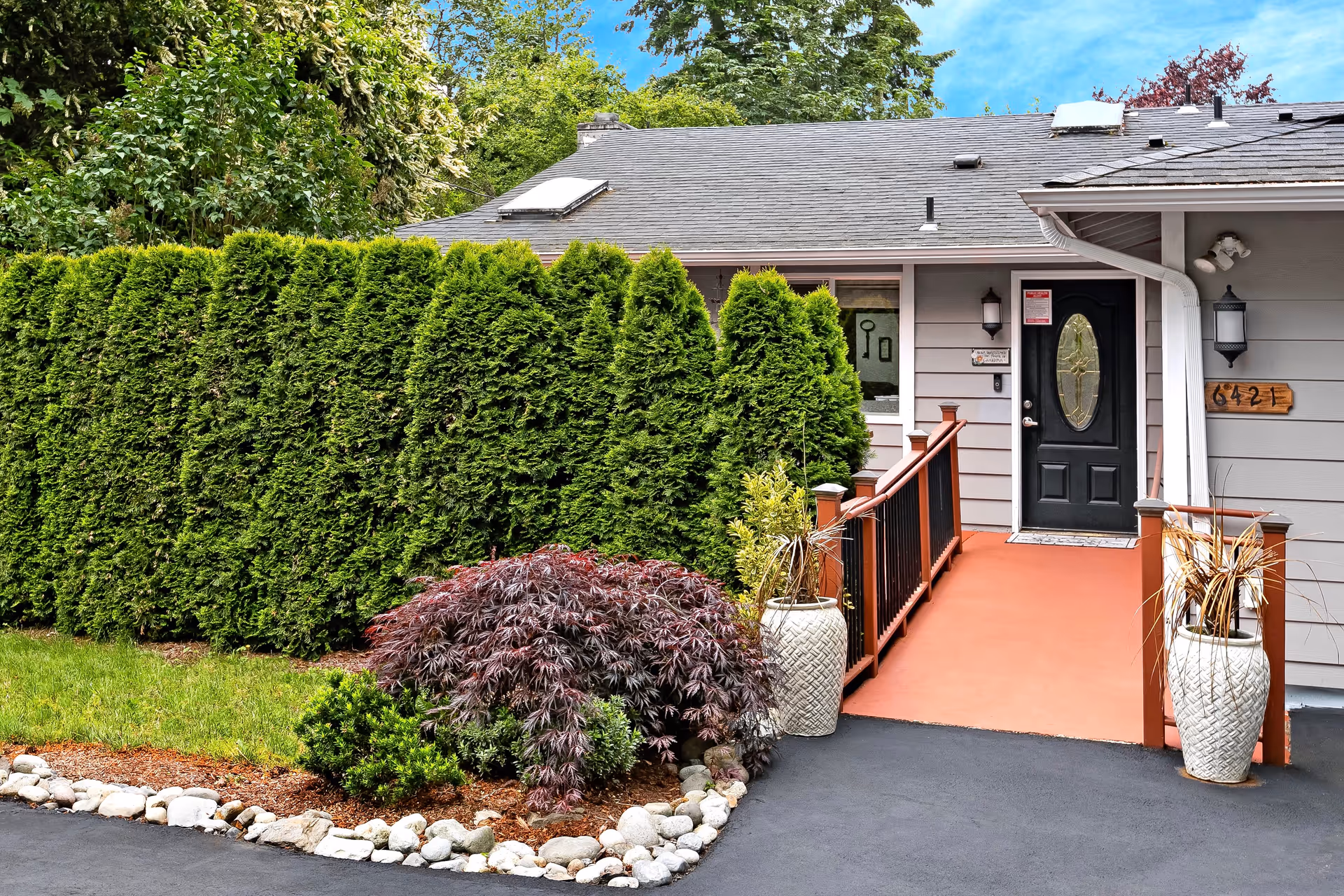Front entrance of a senior care home with a black door, a ramp with red railings, and two large white planters on either side. The house is gray with a shingled roof and surrounded by green bushes and trees. The house number 6421 is displayed on a wooden plaque next to the door.