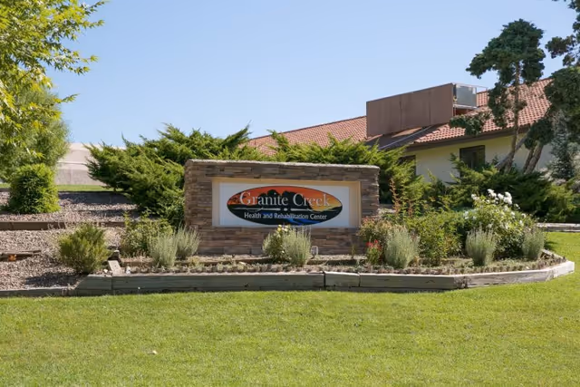 Outdoor view of the Granite Creek Health and Rehabilitation Center sign surrounded by greenery and landscaping with a building and trees in the background under a clear blue sky.