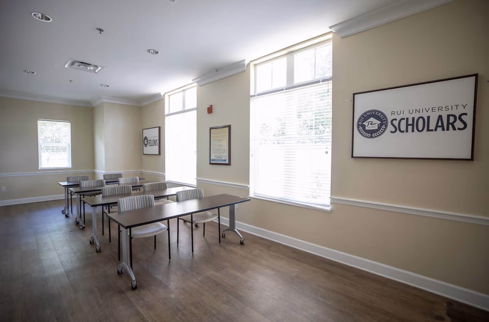 Bright multipurpose activity/meeting room with rows of tables and chairs and a framed "RUI University Scholars" sign on the wall.