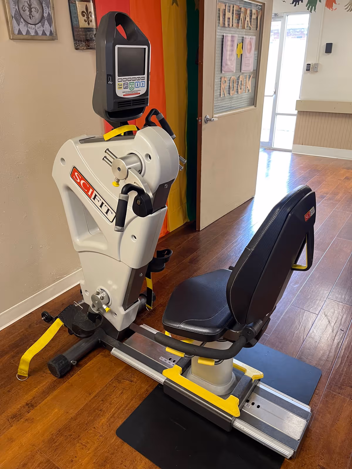 A SciFit seated exercise machine on a mat in a therapy area with wood flooring and a door labeled "Therapy Room".