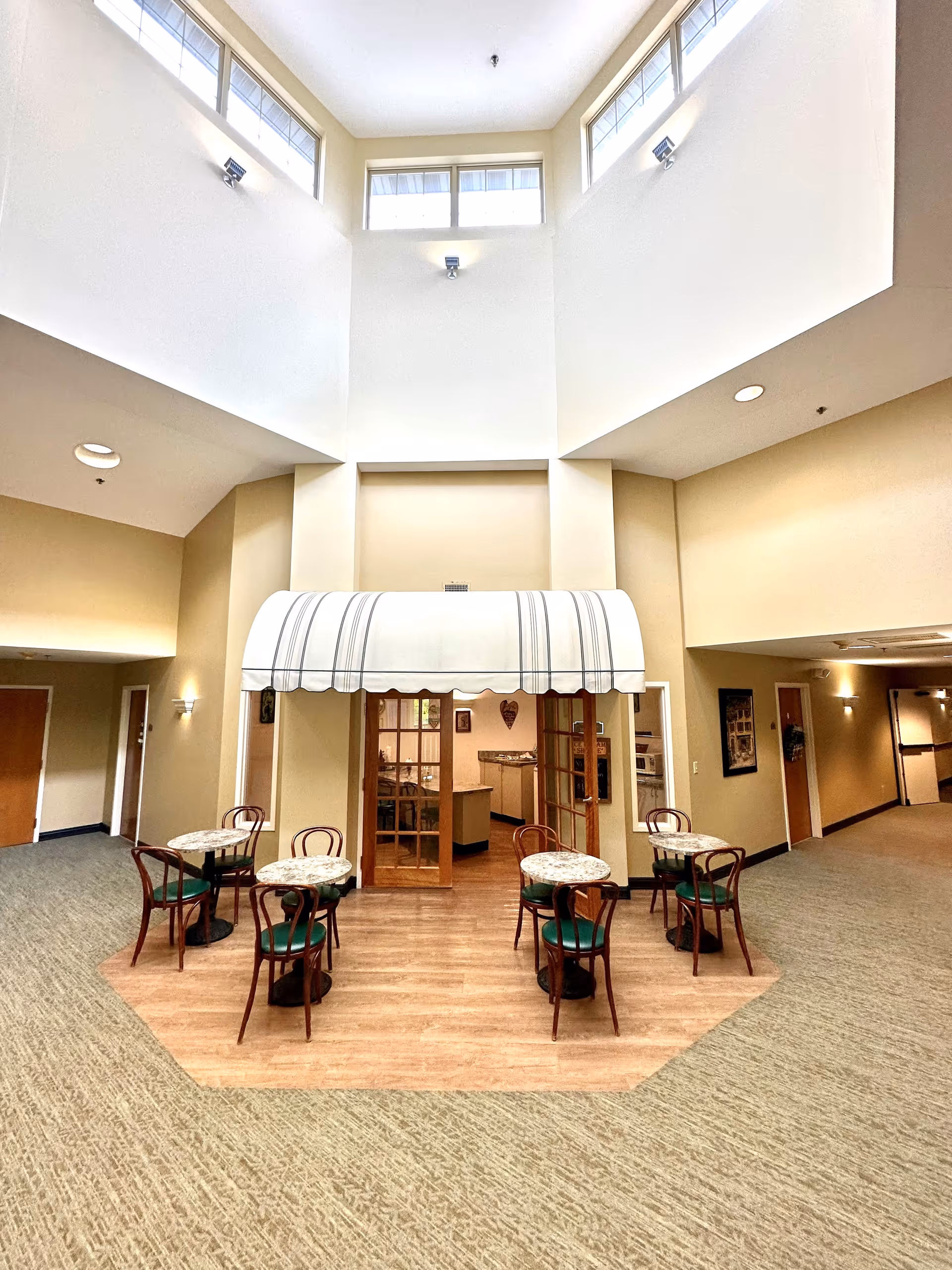 Interior view of a senior living community common area with high ceilings and upper windows. There are four small round tables with green cushioned chairs arranged on a wooden floor section. Behind the tables is a small area with a striped awning and glass double doors leading to a kitchen or dining preparation area. The surrounding floor is carpeted, and the walls are painted beige with some wall decorations and doors visible in the hallway.