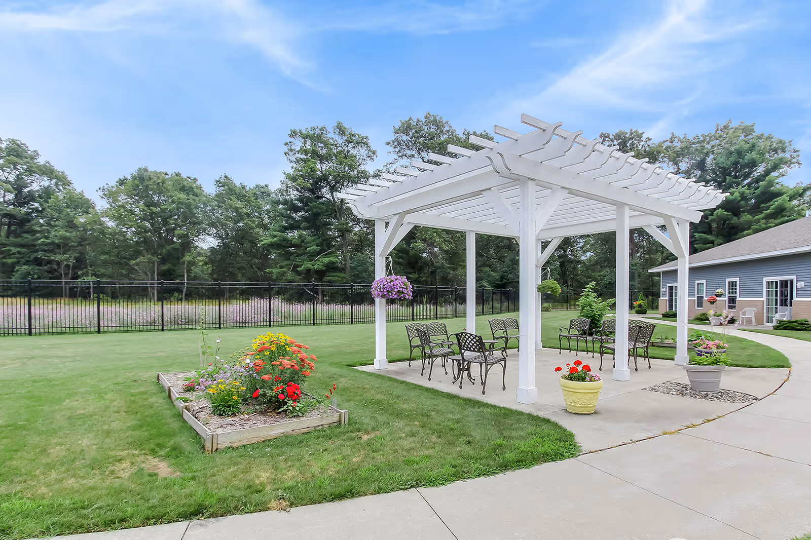 Outdoor seating area with a white pergola structure, metal chairs and tables on a concrete patio surrounded by green grass, flower beds with colorful flowers, and trees in the background under a blue sky.
