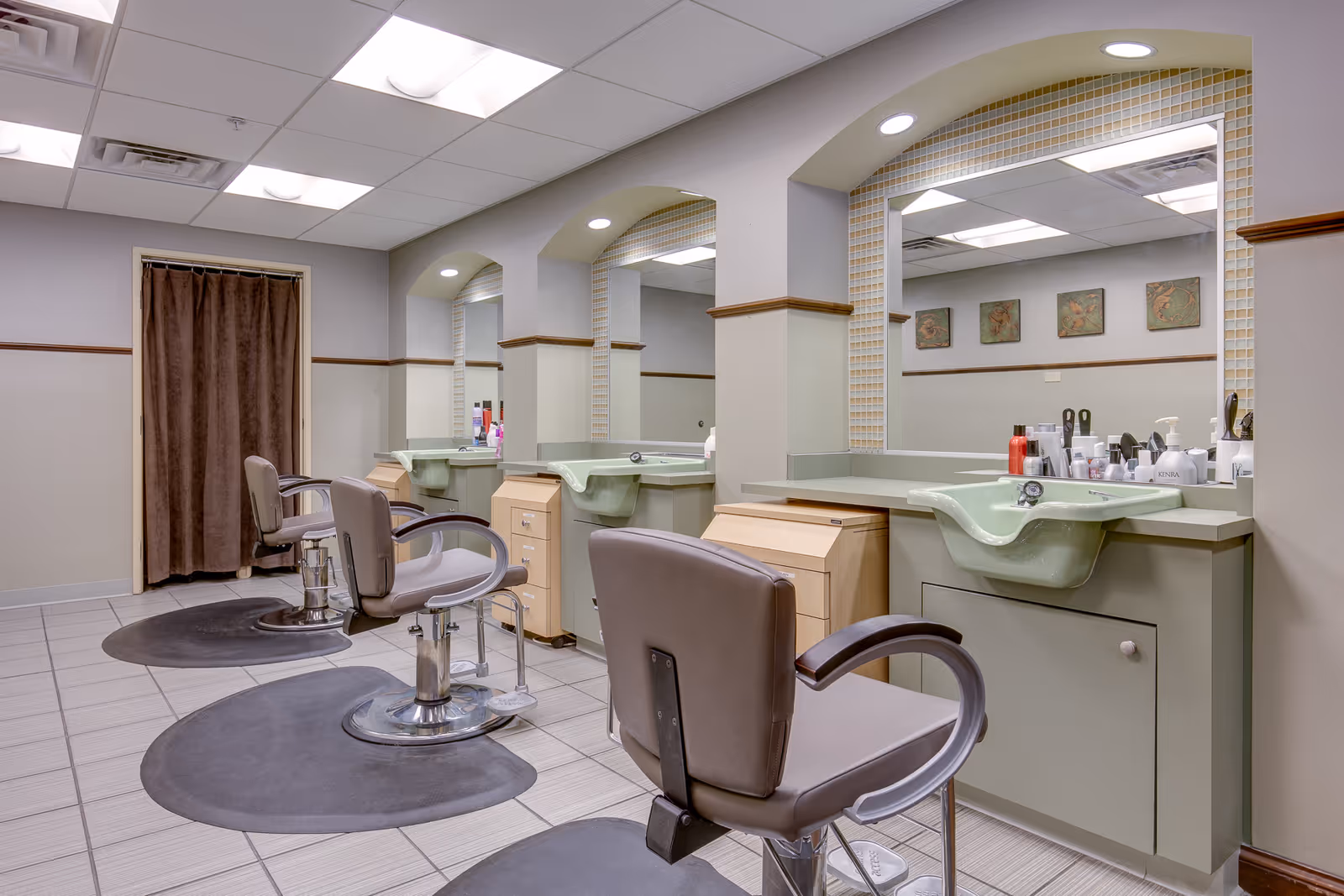 Interior view of a salon area with three styling chairs in front of large mirrors. Each station has a green sink, countertop with various hair care products, and storage cabinets. The room has tiled flooring, a drop ceiling with fluorescent lighting, and a brown curtain covering a doorway.