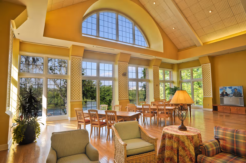 Bright, high‑ceilinged common room with large arched windows, dining tables and lounge seating overlooking trees and a pond.