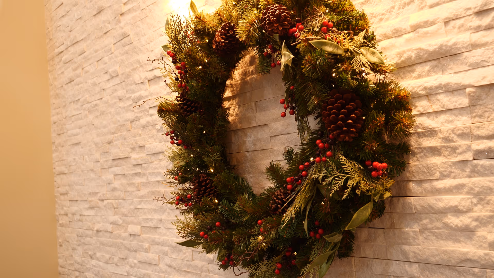 Decorative holiday wreath with pinecones and red berries hanging on a textured stone interior wall.