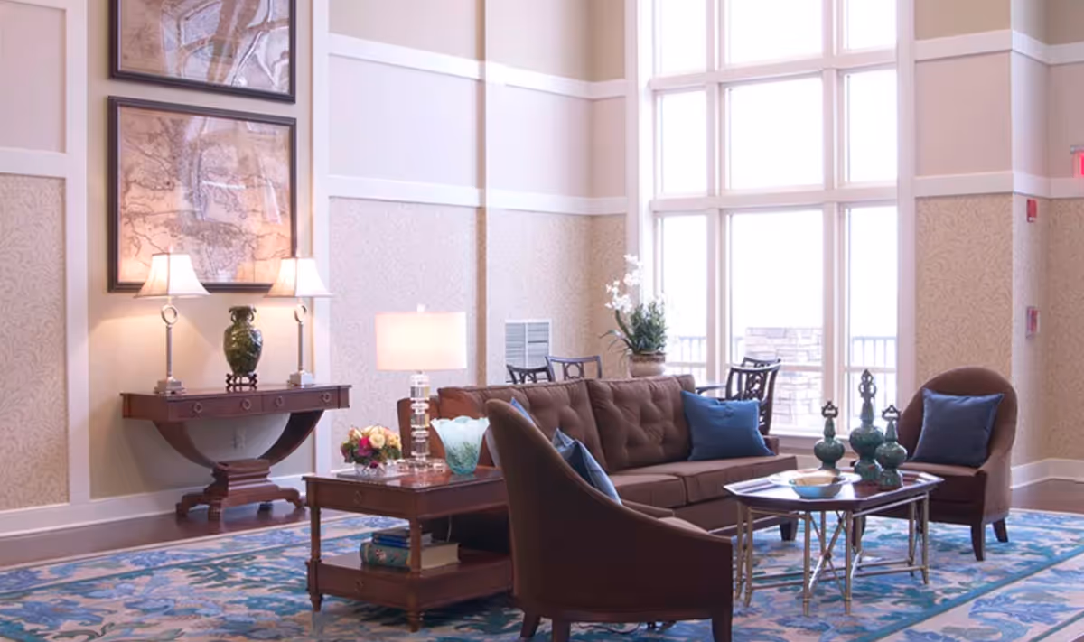 A bright and elegant living room with large windows letting in natural light. The room features a brown tufted sofa and two matching armchairs with blue cushions, a wooden coffee table with decorative vases, a side table with a lamp and flowers, and a console table with two lamps and a green vase. The walls have a patterned wallpaper and two framed maps hang above the console table. A blue and white patterned rug covers the floor.