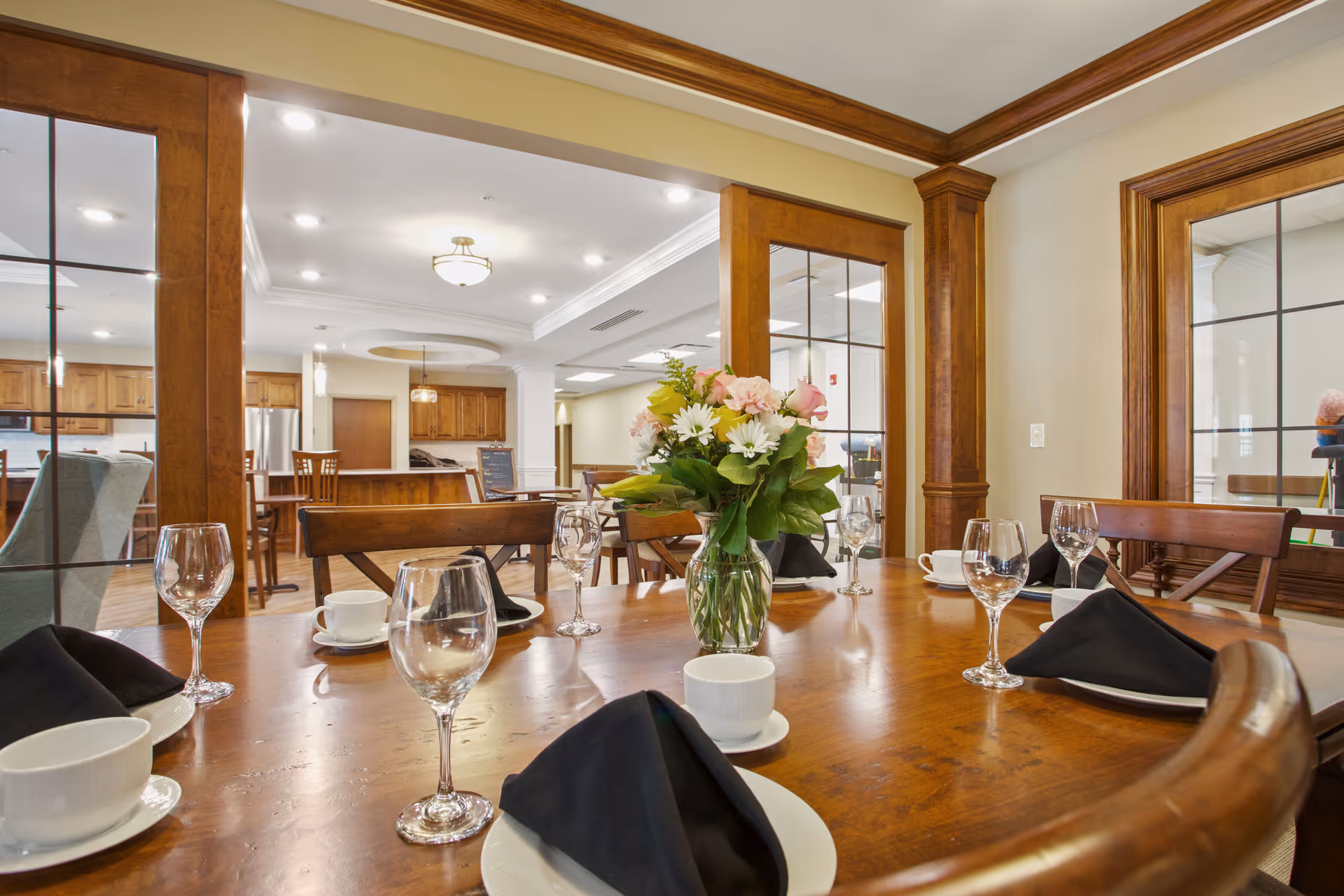 A dining table set with white cups, wine glasses, black napkins on white plates, and a vase of flowers in the center. The room features wooden framed glass doors and windows, with a view into a kitchen area with wooden cabinets and stainless steel appliances.