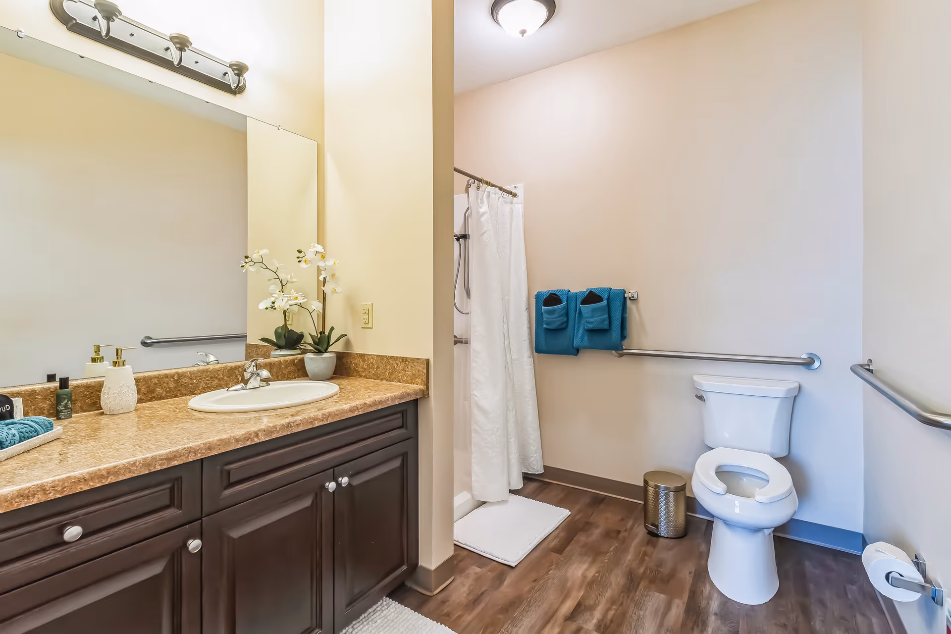 A clean and well-lit bathroom featuring a white toilet with grab bars on the walls, a shower with a white curtain, and a vanity with a brown countertop and dark wood cabinets. The vanity has a sink, soap dispensers, a small potted plant with white flowers, and a large mirror above it. Blue towels hang on a towel rack near the shower.
