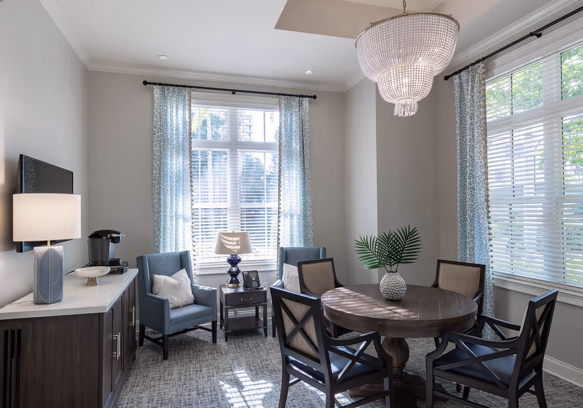 Sunlit seating area with a round wooden dining table and chairs, two upholstered armchairs by large windows, and a sideboard with a lamp and TV.