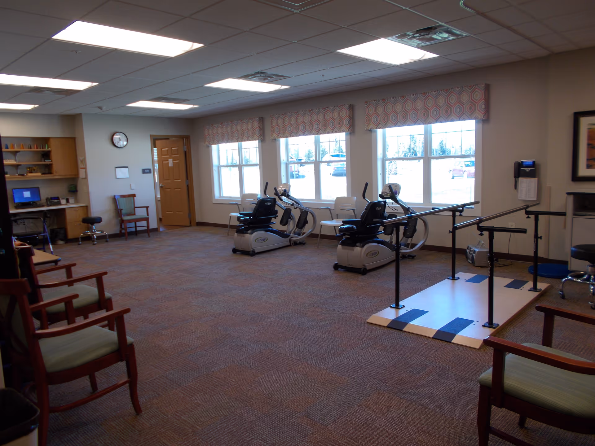 A spacious rehabilitation room with recumbent exercise bikes, parallel walking bars, chairs, and large windows.