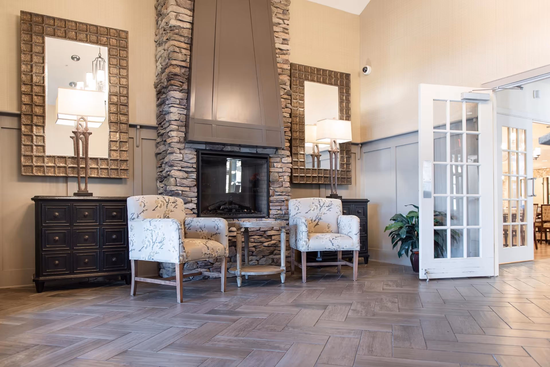 A cozy interior seating area featuring two patterned armchairs placed in front of a stone fireplace with a wooden mantel. On either side of the fireplace are dark wooden cabinets topped with decorative lamps and large framed mirrors. To the right, white French doors open into another room with visible dining furniture and a potted plant nearby.