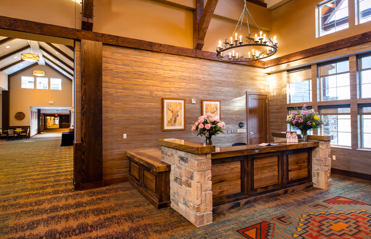 Reception area in a senior living facility with a wooden desk featuring stone accents, flower arrangements on the desk, framed artwork on the wall, large windows letting in natural light, and a chandelier hanging from a high ceiling with exposed wooden beams. A hallway with seating and lighting fixtures is visible in the background.