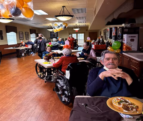 Residents in wheelchairs eating and socializing in a decorated senior living dining room.