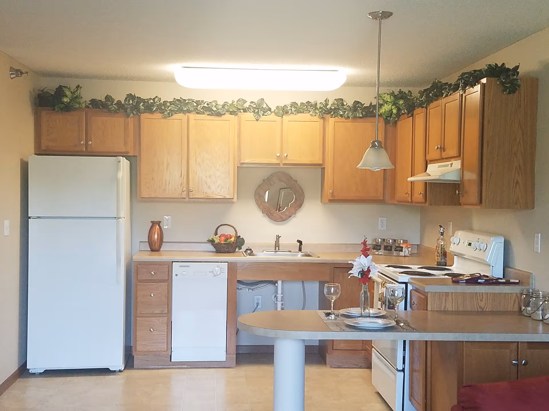 A kitchen with wooden cabinets, a white refrigerator, dishwasher, and stove. There is a countertop with a small dining area set with plates, glasses, and a vase with flowers. Greenery decorates the top of the cabinets.