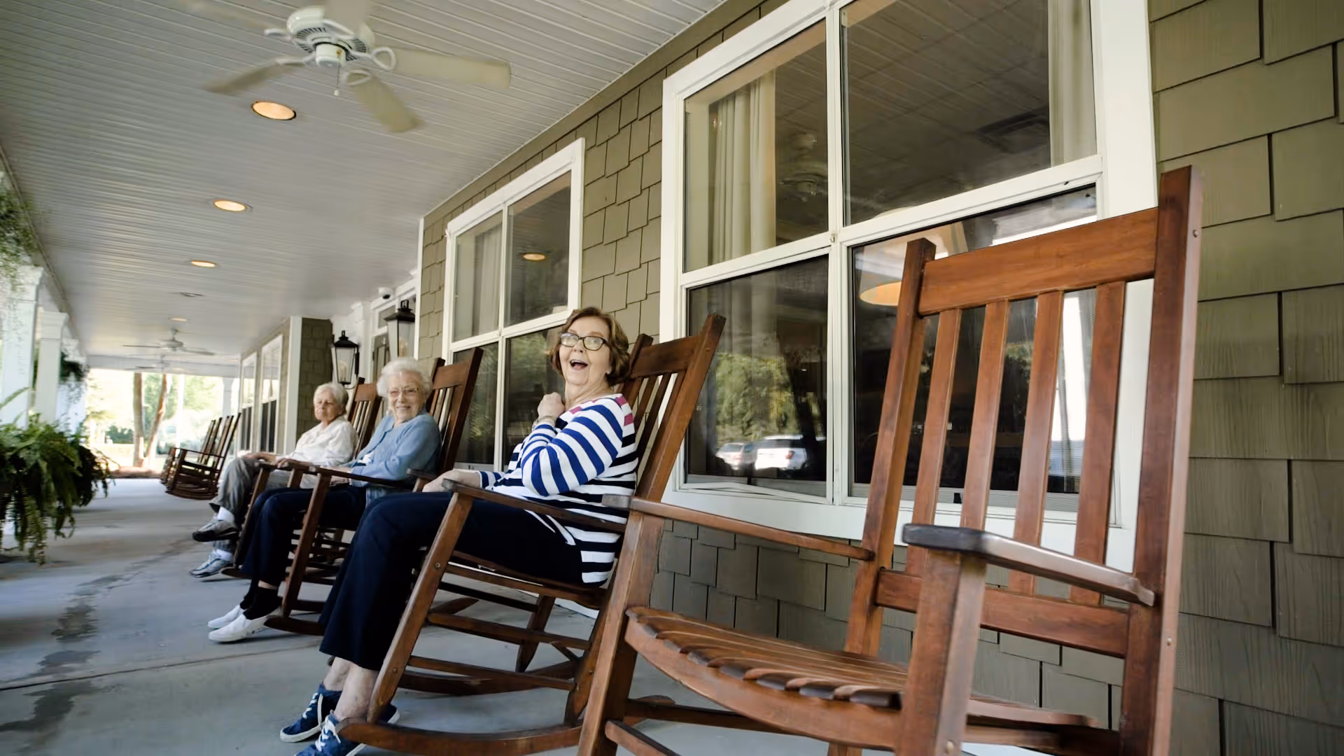 Three elderly women sitting and smiling on wooden rocking chairs on a covered porch with green siding and large windows, with ceiling fans overhead and plants hanging nearby.