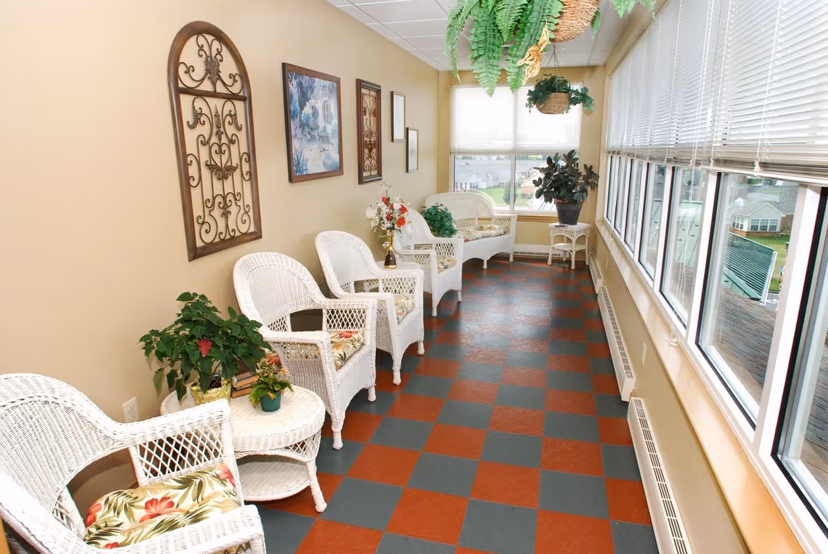A sunroom with large windows on one side, featuring white wicker chairs and tables with floral cushions and potted plants. The floor has a red and gray checkered pattern, and the walls are decorated with framed artwork and metal wall decor. Hanging plants are suspended from the ceiling.