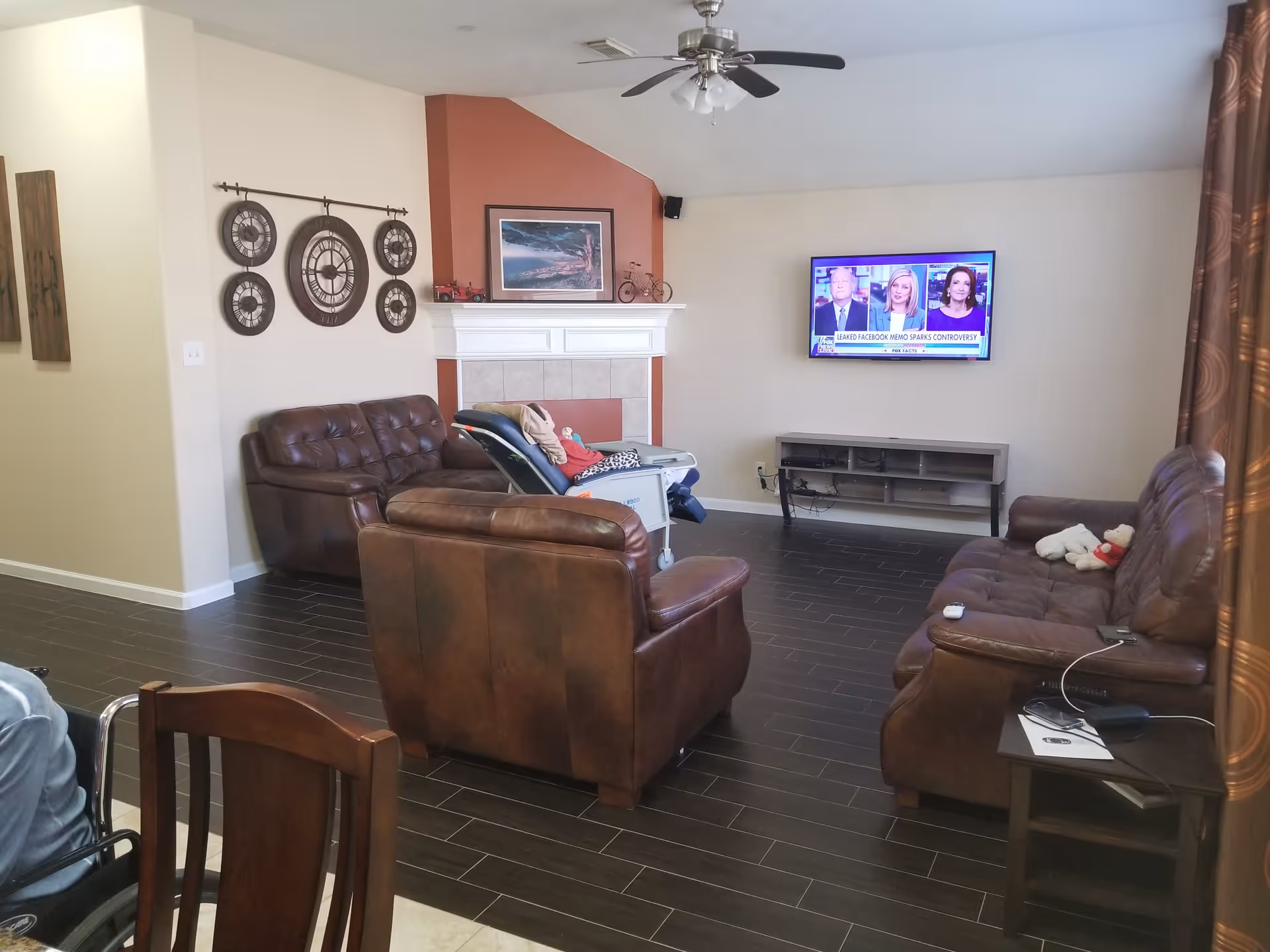 A cozy living room with brown leather sofas arranged around a fireplace with a red accent wall. A flat-screen TV is mounted on the wall opposite the sofas, showing a news program. Decorative wall clocks hang on one wall, and a ceiling fan with lights is installed overhead. There is a small side table with some items on it and a wheelchair partially visible in the foreground.