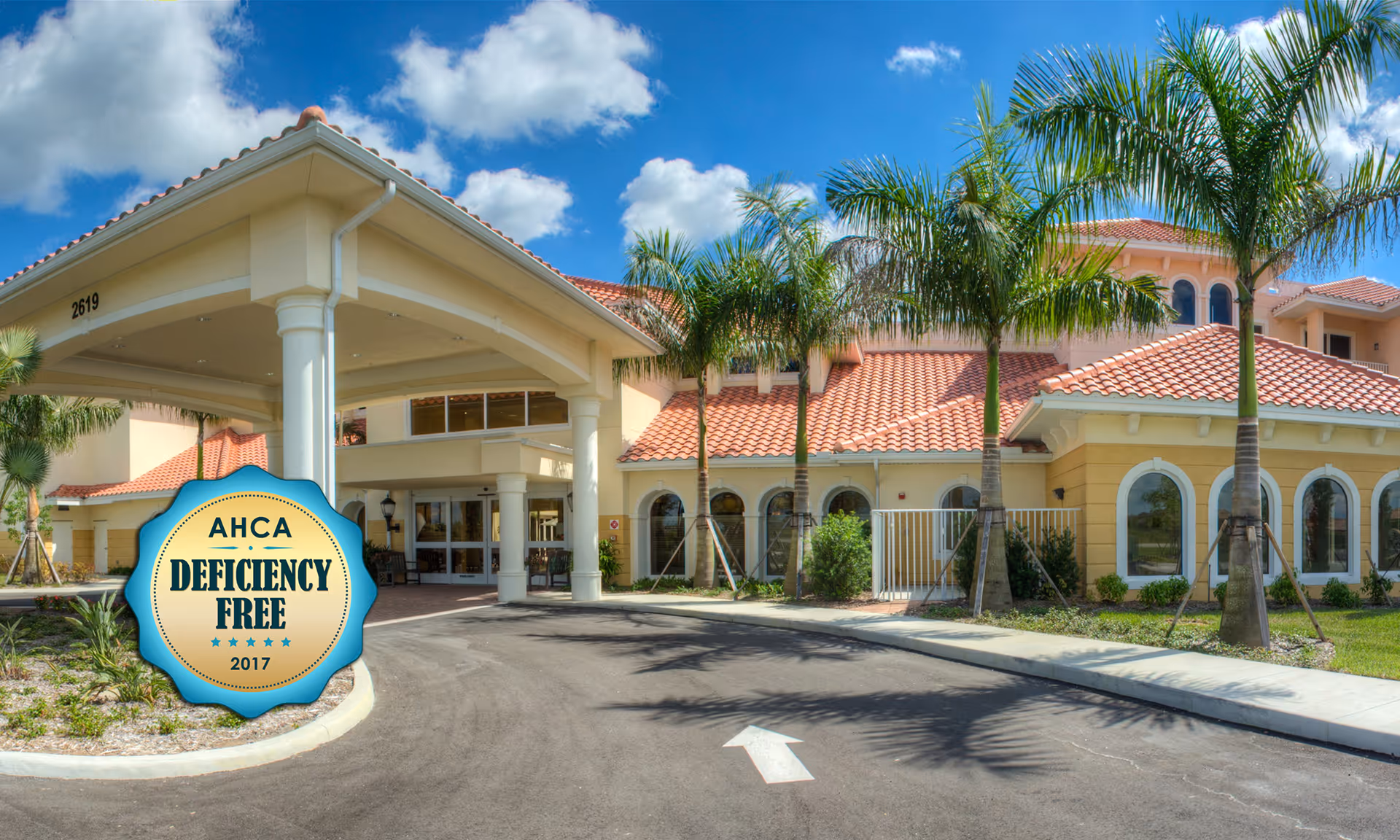 Exterior view of Discovery Village At The Forum senior living facility with a covered entrance, palm trees, and a clear blue sky with some clouds.