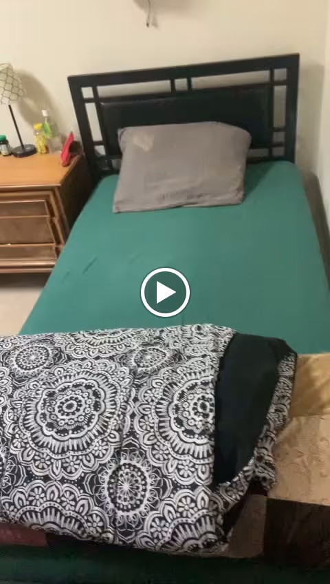 A single bed with a black metal headboard, a gray pillow, and a green fitted sheet. A patterned black and white blanket is folded at the foot of the bed. To the left of the bed is a wooden nightstand with a lamp, a bottle of hand sanitizer, and other small items.