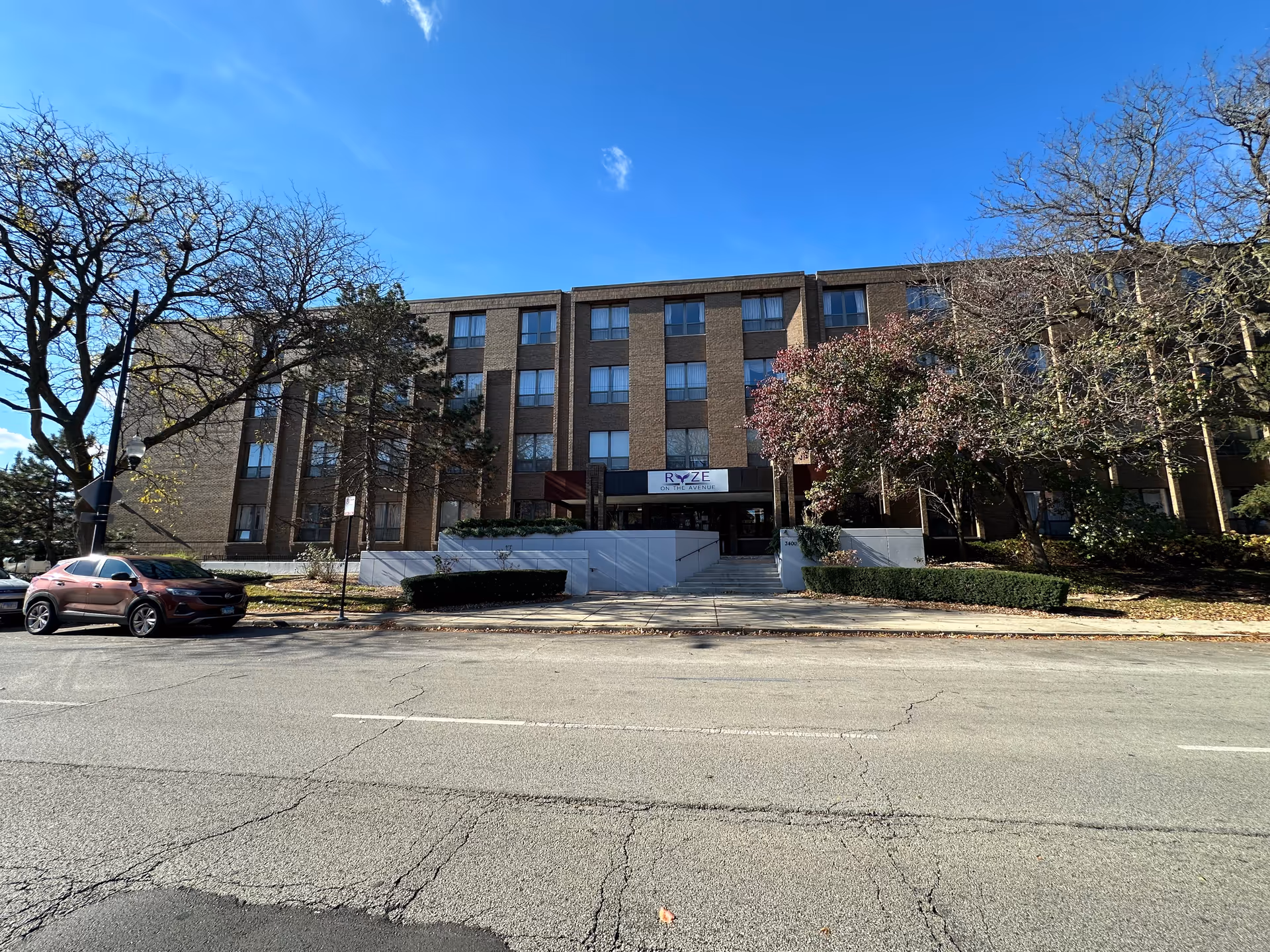 Front facade of a four-story brick senior living building with a sign reading "RYZE" above the main entrance, flanked by trees and a parked car.