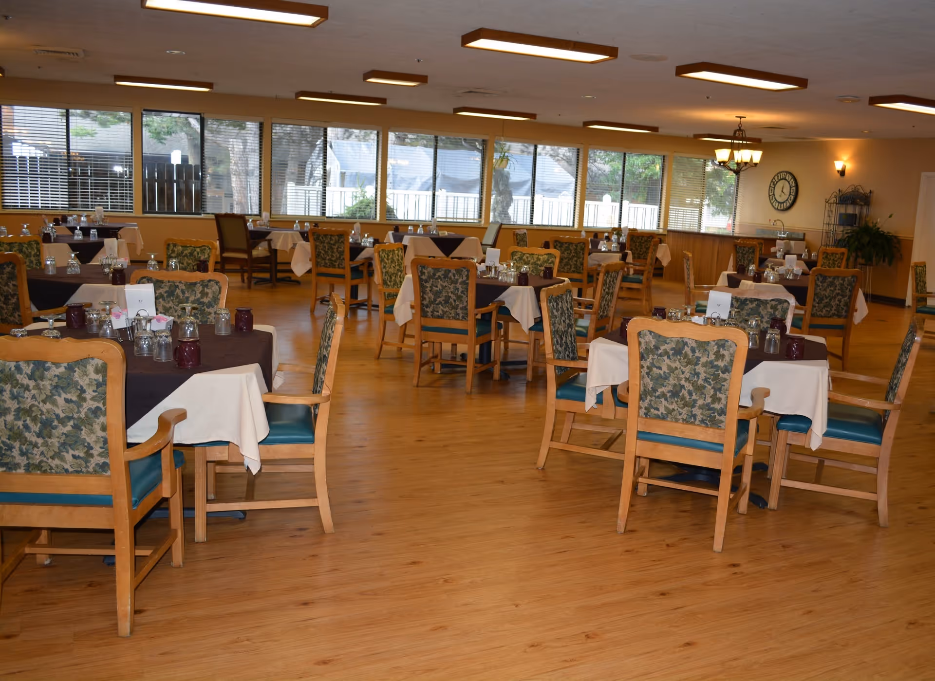 A spacious dining room with multiple wooden tables and chairs arranged neatly. Each table is covered with a white tablecloth and a dark runner, set with glasses and small decorative items. Large windows line the far wall, allowing natural light to fill the room. The floor is wooden, and the ceiling has rectangular light fixtures. A clock and some plants are visible on the walls and corners.