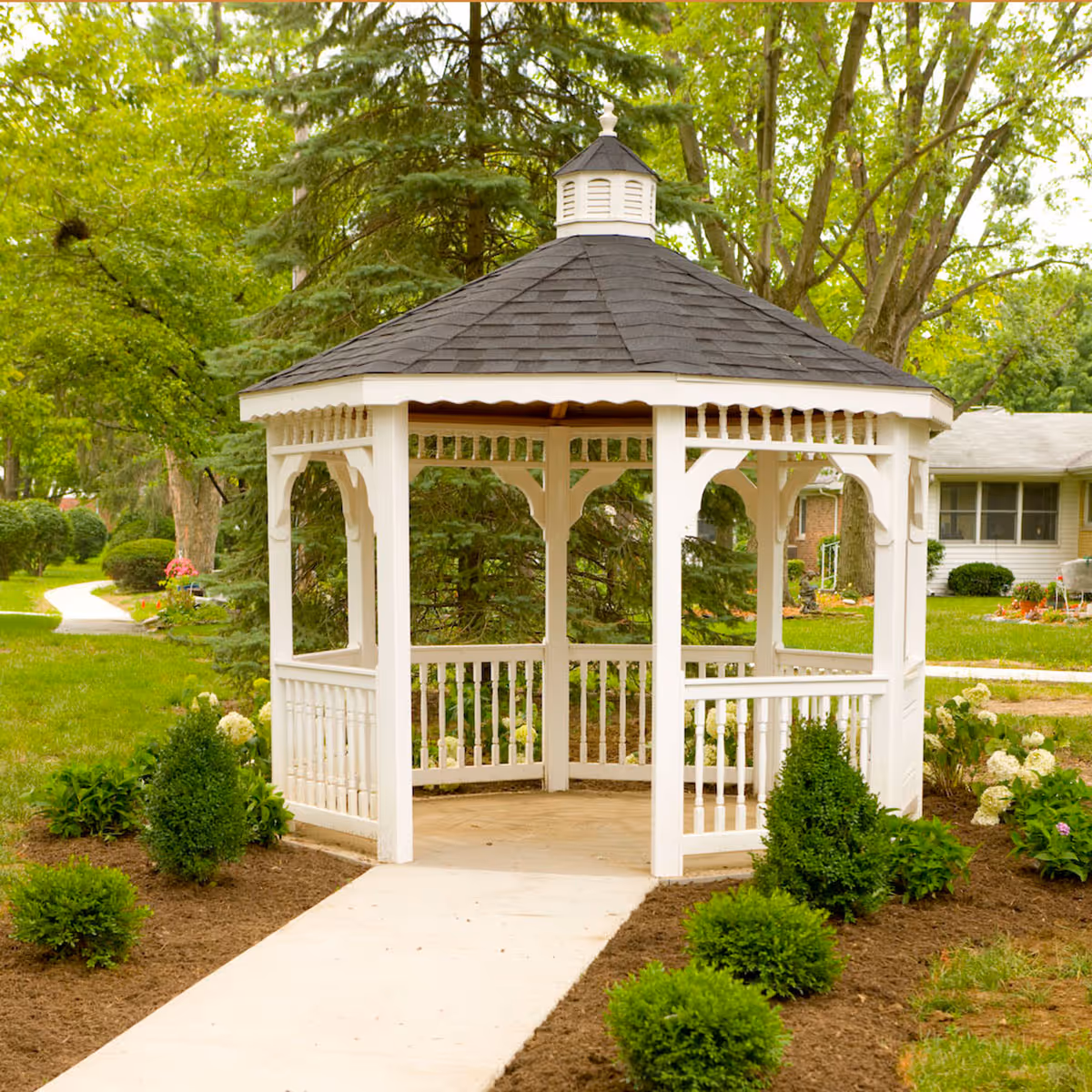 A white wooden gazebo with a dark shingled roof situated in a garden area with green shrubs and trees surrounding it. A concrete pathway leads up to the gazebo, and a house is visible in the background.