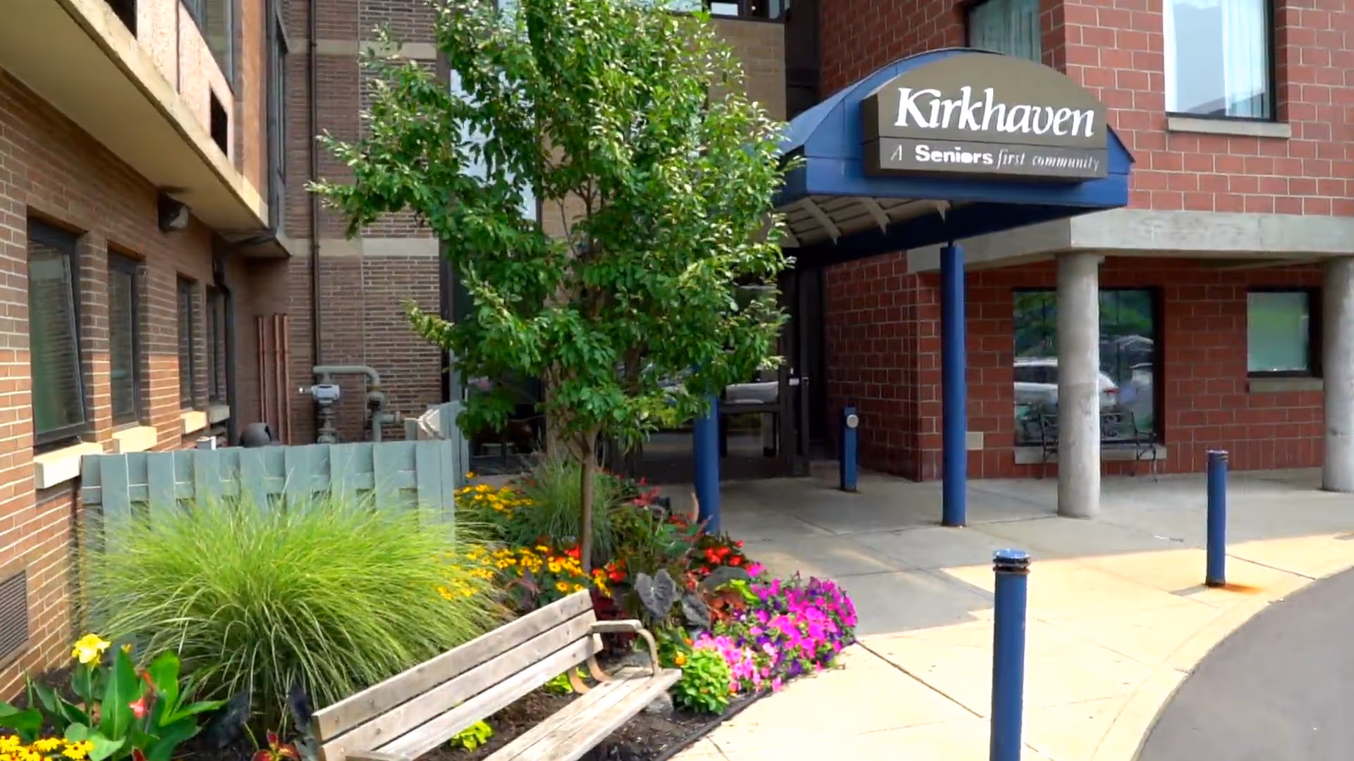 Entrance to Kirkhaven senior living facility with a covered walkway, a wooden bench, and a landscaped garden featuring green shrubs, colorful flowers, and a small tree. The building exterior is made of red brick with windows and concrete pillars.