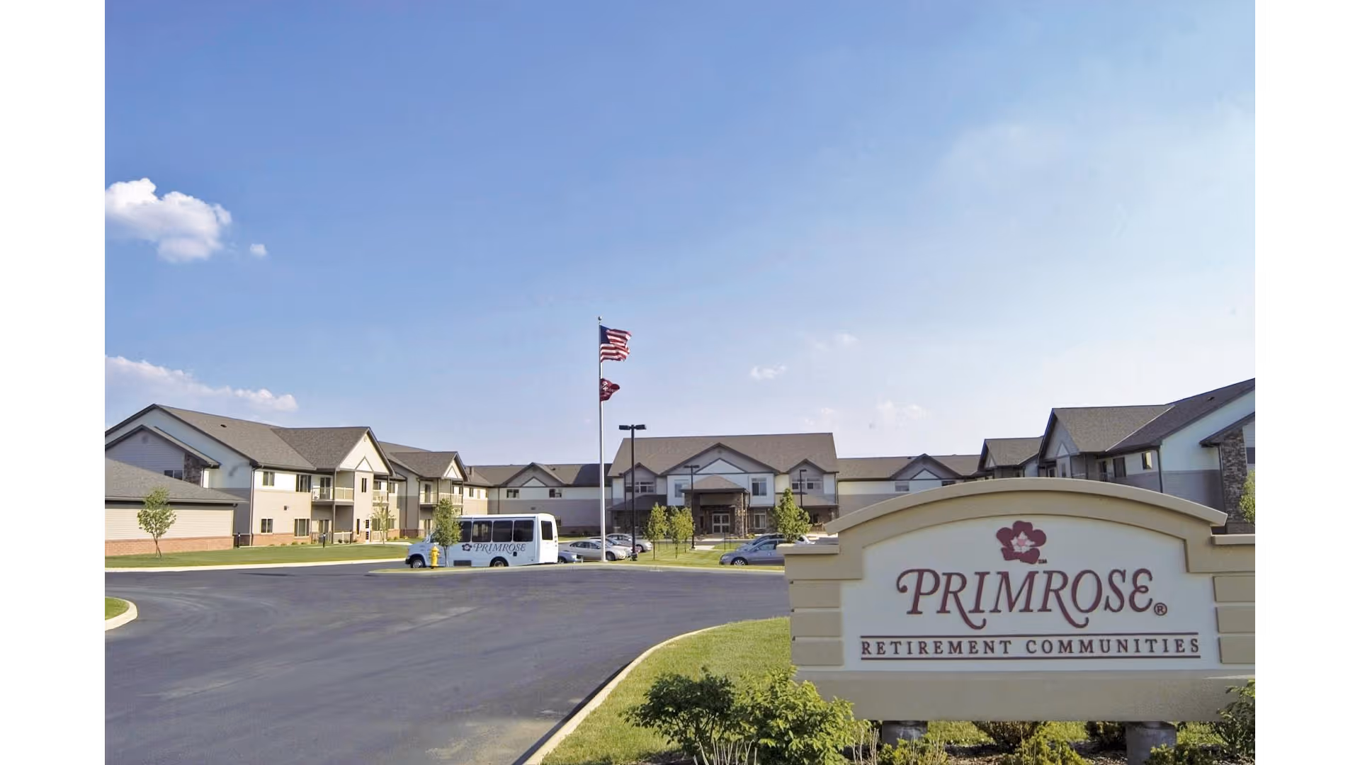 Exterior view of Primrose Retirement Community of Marion showing a large building with multiple windows and a peaked roof under a clear blue sky. There is a parking lot with a white shuttle bus and several cars. In the foreground, there is a sign with the Primrose Retirement Communities logo and name. Two flagpoles with American and state flags are visible near the building entrance.