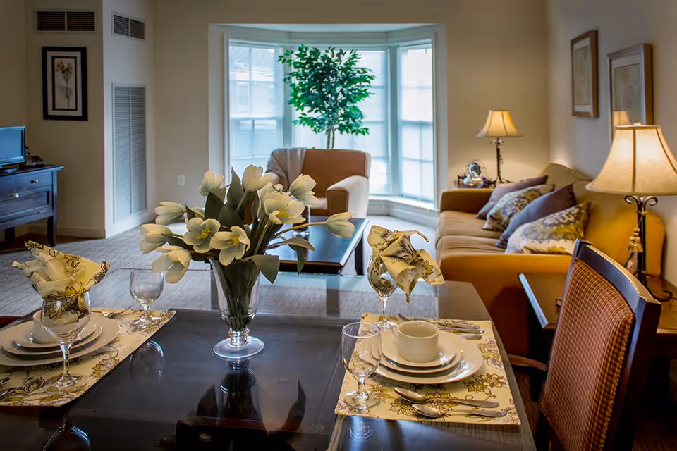 A cozy living and dining area in an assisted living community featuring a dining table set for two with plates, glasses, and floral napkins. A vase with white flowers is centered on the table. In the background, there is a comfortable seating area with a sofa, armchair, decorative pillows, a coffee table, a floor lamp, and a large window with a potted plant.