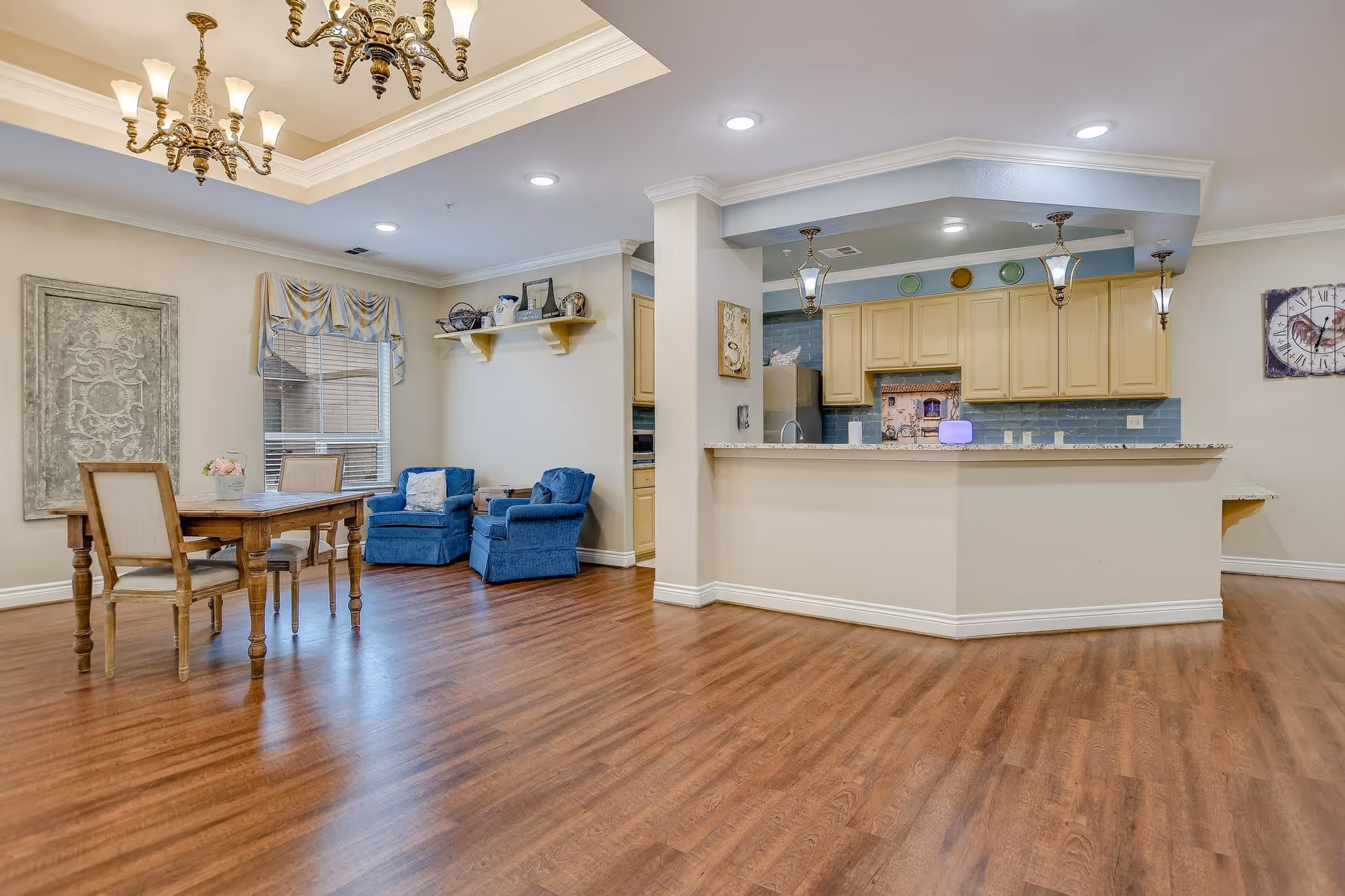 Interior view of a memory care facility showing a dining area with a wooden table and four chairs, two blue armchairs near a window with a decorative valance, and a kitchen with yellow cabinets, a granite countertop, and hanging pendant lights. The floor is wooden, and the ceiling has recessed lighting and a chandelier.