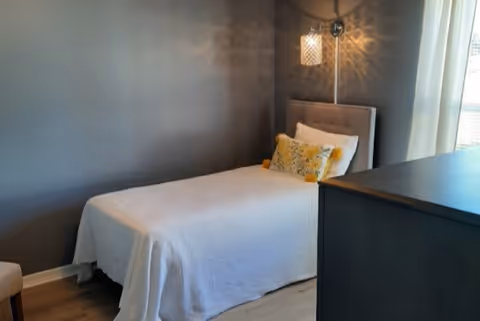 A small bedroom with a single bed covered in a white blanket, a decorative yellow pillow, a wall-mounted lamp with a patterned shade casting light on the gray wall, and a dark dresser near a window with sheer curtains.
