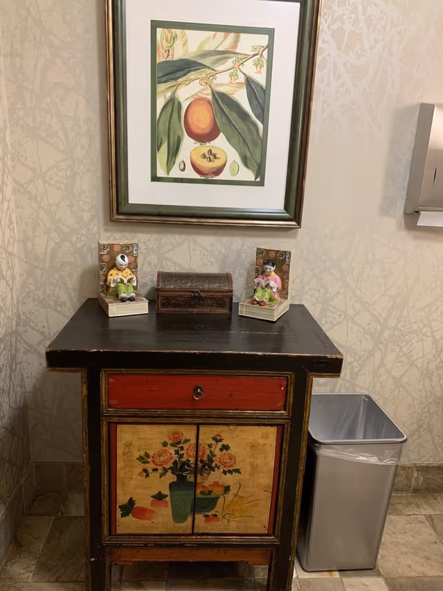 A decorative wooden cabinet with a red drawer and floral painted doors, topped with two small figurines and a small wooden chest. Above the cabinet hangs a framed botanical print of a fruit and leaves. To the right of the cabinet is a silver trash bin and a wall-mounted paper towel dispenser. The walls have a subtle patterned wallpaper.