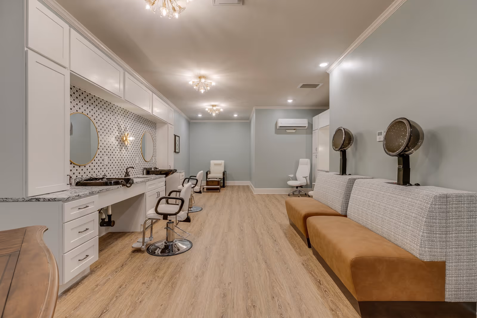 Interior view of a modern salon area with light wood flooring, white cabinetry with marble countertops, two round mirrors, and salon chairs. On the right side, there are two vintage-style hair dryers mounted above a cushioned bench. The walls are painted light blue, and the ceiling has decorative light fixtures.