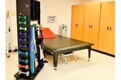 Physical therapy or rehabilitation room with a black treatment table, a rack holding colorful dumbbells, exercise bands, and medical equipment mounted on the wall. Wooden storage cabinets are visible in the background.