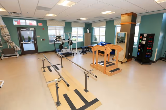 A spacious physical therapy room with parallel bars for walking exercises, a wooden staircase with handrails for step training, exercise equipment including a treadmill and weights, and large windows letting in natural light.