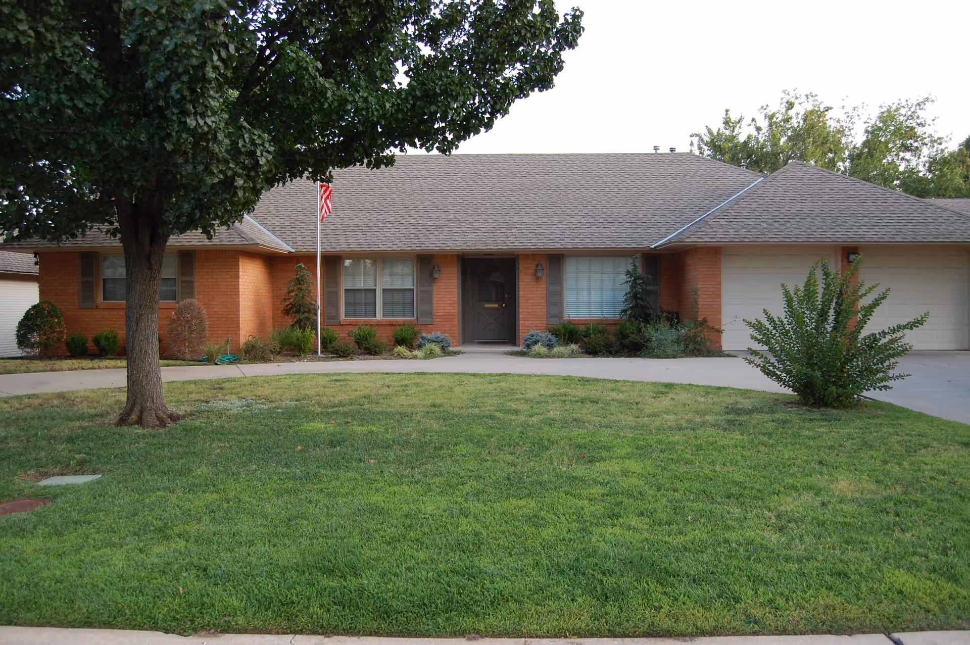 Front exterior view of a single-story brick house with a gray shingled roof, two large windows with shutters, a central front door, a two-car garage on the right, a green lawn with a tree on the left, and an American flag on a flagpole near the front entrance.