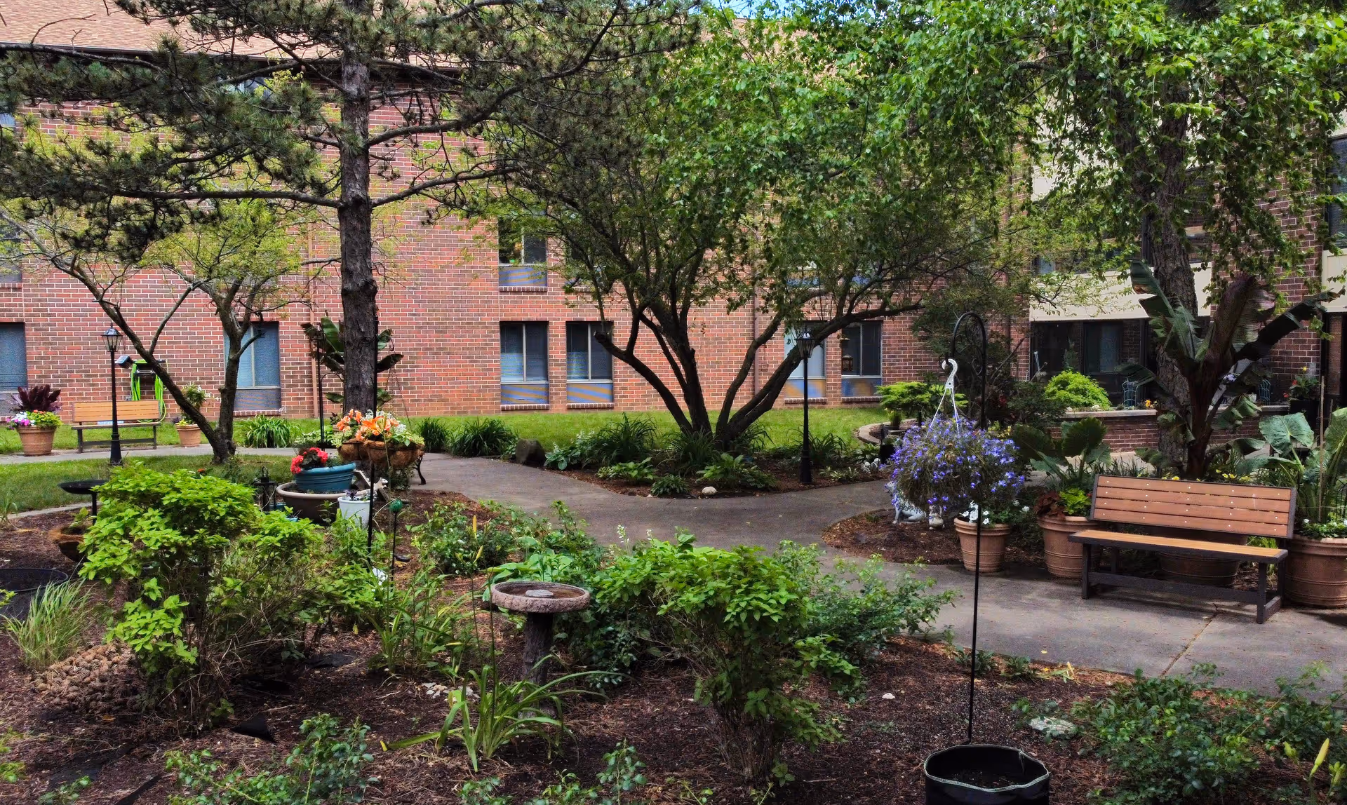 A peaceful outdoor garden area at Independence Village featuring a variety of green plants, trees, and flowers. There are paved walkways, a wooden bench, potted plants, and a birdbath. The background shows a brick building with several windows.