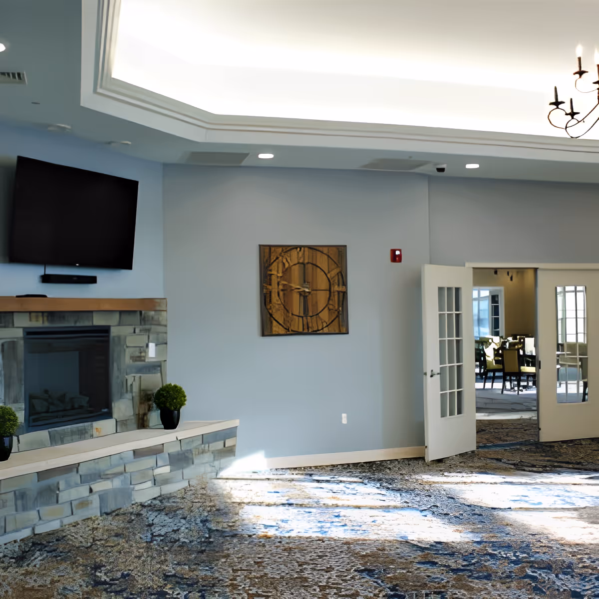 A bright and spacious senior living common area with a stone fireplace, a mounted flat-screen TV above it, two small potted plants on the fireplace ledge, a large decorative wooden clock on the wall, and double glass doors leading to a dining area with tables and chairs.