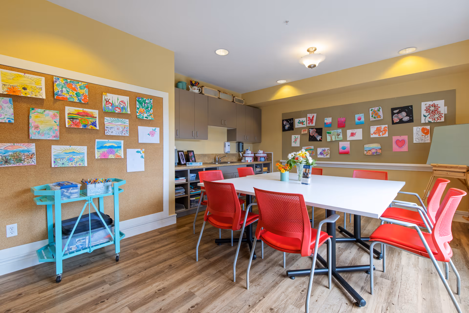 Bright activity room with a white table surrounded by red chairs, corkboards covered in colorful artwork, and cabinets and supplies along the back wall.